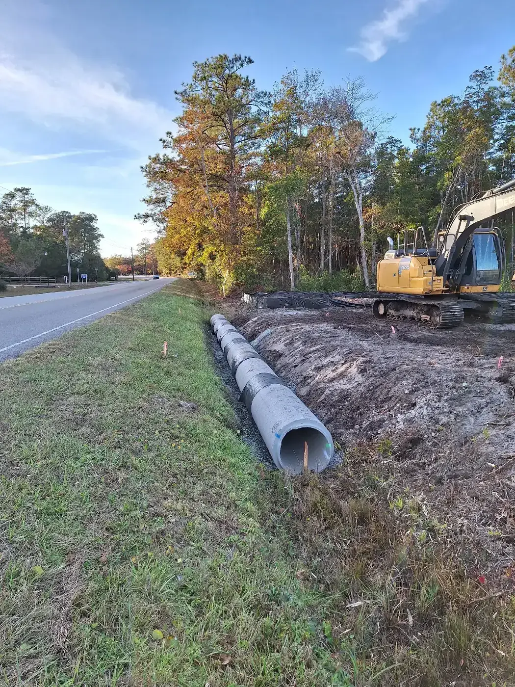 Concrete culvert pipe laid along a roadside ditch beside a two-lane road, with an excavator on the shoulder, in southeastern North Carolina
